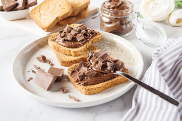 Spreading sweet chocolate cream, cashew, chocolate chunks onto toast on table.