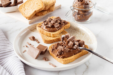 Spreading sweet chocolate cream, cashew, chocolate chunks onto toast on table.