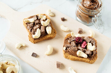 Spreading sweet chocolate cream, cashew, chocolate chunks onto toast on table.