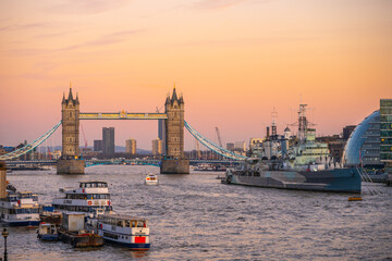 Obraz premium Tower Bridge stands majestically over the Thames River as HMS Belfast is docked nearby. The evening sky adds a warm glow to the iconic London skyline, showcasing the city's historical charm.