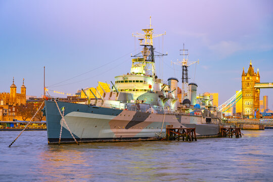 HMS Belfast is docked along the Thames River, illuminated by evening light. The famous Tower Bridge stands majestically in the background, creating a stunning urban view.