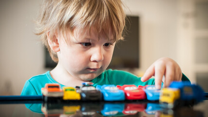 A young child examines and organizes a collection of toy cars on a reflective surface, showcasing curiosity and concentration in a bright indoor setting