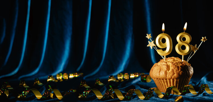 Number 98 gold celebration candle and small stars in sweet cupcake on bright blue velvet background. Concept of celebrating birthday, anniversary. Banner.