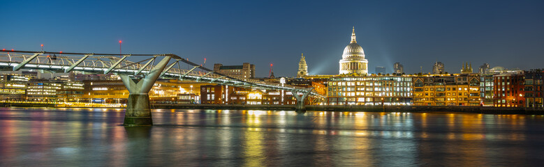 Fototapeta premium Twinkling lights illuminate Millenium Bridge and St. Paul's Cathedral as dusk settles over the River Thames, creating a captivating view of London in the evening.