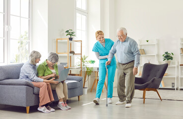 Group of happy elderly people in the living room of a nursing home. A nurse helps an older man walk...