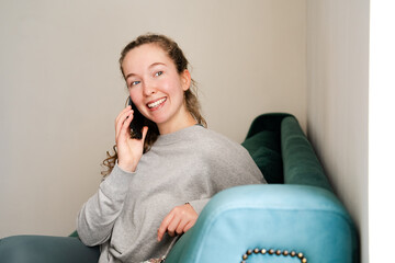 Beautiful positive young woman talking on the phone sitting on a couch at home. Happy girl speaking...