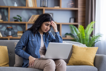 Naklejka premium Upset young Indian woman sitting on the couch at home, holding a laptop on her lap and looking worriedly at the screen while holding her head in her hand
