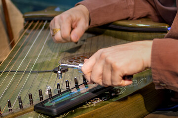 Close-up of man's hands tuning traditional stringed instrument using tuning tool and smartphone...