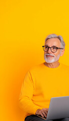 Senior man with glasses, yellow sweater, using laptop against yellow background