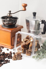 vintage coffee grinder and moka pot sit beside container of coffee ice cubes, surrounded by scattered coffee beans and eucalyptus leaves, creating cozy coffee themed scene