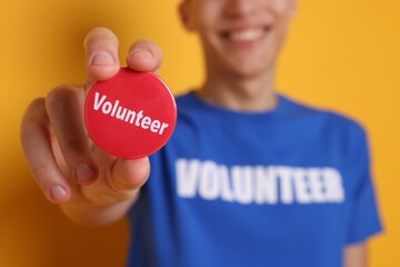 Man holding button badge with word Volunteer on orange background, closeup