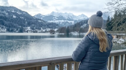 Fototapeta premium Woman in a cozy winter outfit gazes over a serene lake surrounded by snow-capped mountains and trees