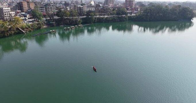 aerial shot reveals a single small canoe with occupants as they navigate on Phewa Lake. The lake stretches towards the far shore, which is immediately adjacent to the densely populated city of Pokhara