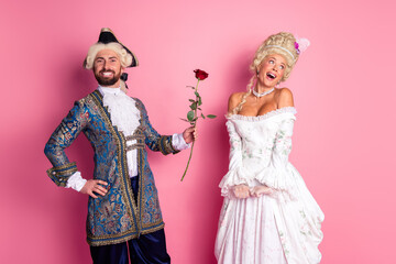 Elegant couple in historical royal costumes with a vibrant pink backdrop showcasing a romantic scene holding a rose