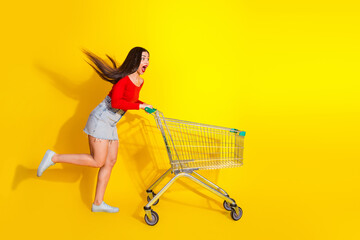 Young woman playfully pushing a shopping cart on a vibrant yellow background, expressing joy and...