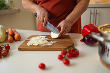 A person slices onion on a wooden cutting board in a bright kitchen. Various fresh vegetables,...