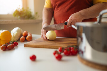 A woman in a yellow shirt and apron is peeling an onion in the kitchen. She is preparing ingredients for a meal while surrounded by fresh vegetables like carrots, tomatoes, and bell peppers