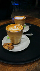 Portrait of love-shaped cappuccino coffee in a glass cup with white saucer and biscuits on a wooden table background.