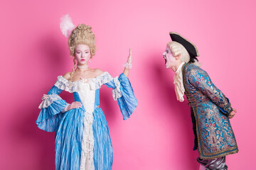 Elegant couple in baroque costumes posing on pink background, embodying classic style with a blend of humor and drama