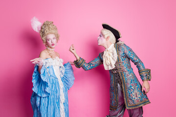 Elegant couple in vintage costumes posing against pink background at themed carnival event,...