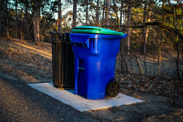 two outdoor trash cans, one black and one blue, sitting on a concrete pad in a wooded park...