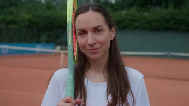 young female tennis player with long brown hair standing on clay court, holding racket and looking confidently at camera, sportswoman in white sportswear during outdoor practice session