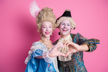 Young couple in historic costumes making a heart shape with hands on a vibrant pink background