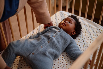 High angle shot of Black mother putting sleeping baby girl dressed in knitted jumpsuit into crib...