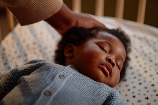 Close up on peaceful face of Black baby girl sleeping in crib while mothers hand gently caressing kids hair in childrens bedroom