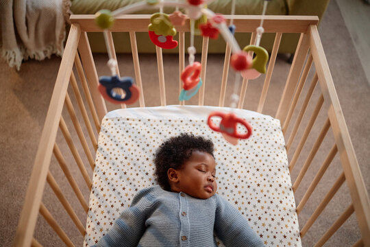 Cropped shot of little Black girl dressed in blue knitted jumpsuit sleeping on starry blanket in baby crib with musical mobiles, hanging above child in home setting - Powered by Adobe