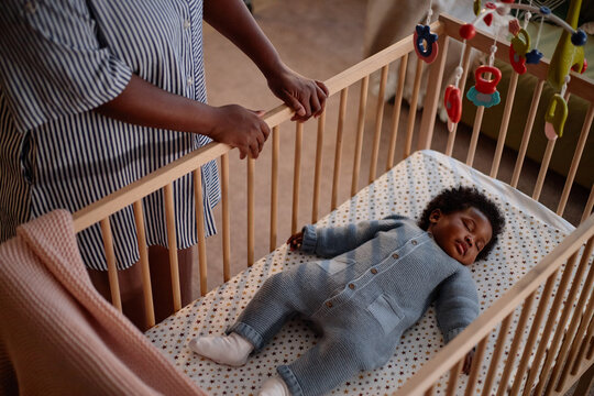 High angle shot of little Black child dressed in knitted jumpsuit of blue color serenely sleeping in cozy wooden crib with baby mobiles hanging above, while mother checking on daughter in bedroom