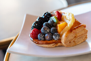 A fruit-filled French puff pastry on a white square plate. The cream and custard-filled dessert has strawberries, raspberries, blueberries, beaches, kiwi, blackberries and is topped with a sugar glaze