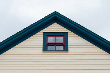 An upward view of a cloudy grey sky and an exterior gable end of a pale yellow wooden house with a small double hung window. The trim around the eave and window is dark green with deep red trim.