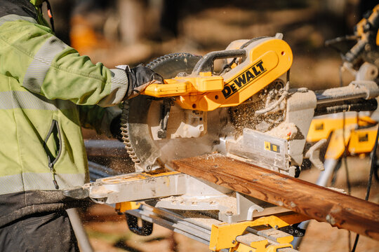 Worker cutting a wooden plank with a DeWalt miter saw outdoors, generating sawdust in sunlight at a construction site..