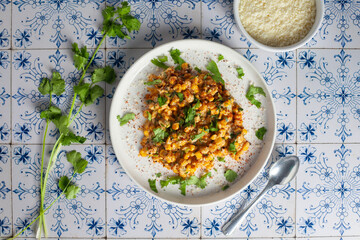 Esquites on a white plate on a blue tile with cilantro, cotija, and a spoon.