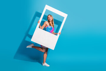 Beautiful happy woman posing with a frame against bright blue background showcasing summer fashion...