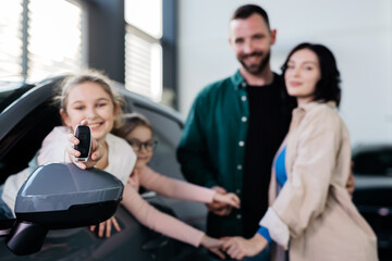 Little girl holding the keys to a new car sitting in the showroom, joyful family in the background hugging each other. Concept of expensive purchase of a new car.