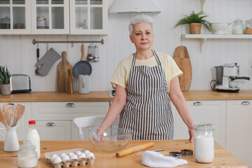 Happy senior woman cooking in kitchen. Stylish older mature gray haired lady grandmother knead...