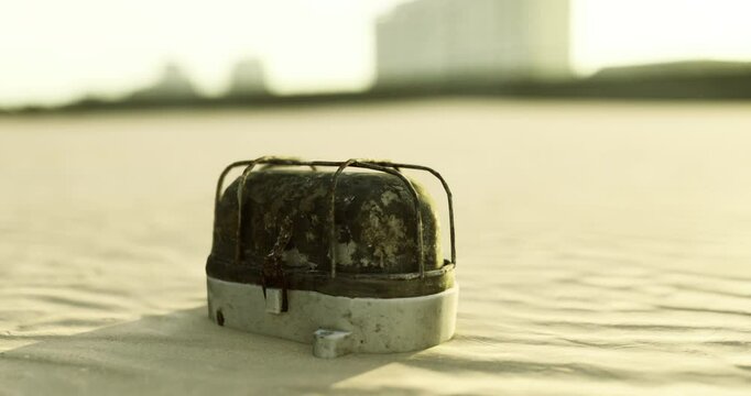 An old camping stove sits on a sandy surface, partially covered in dirt. The sun sets in the background, casting warm light over the scene, creating a serene atmosphere.