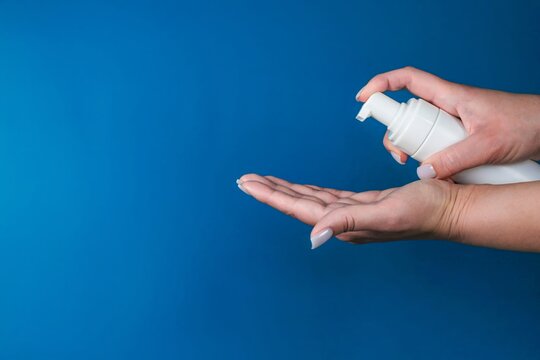 Close-up of two hands, one of them holding a white bottle with a dispenser over the palm of the other hand on a blue background with space for text. The concept of foam cleansers, skin cleansers. High