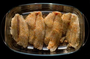 Raw seasoned fish, Nile Tilapia species, in a glass ovenproof dish, in the form of a rectangle. Prepared for baking. Transparent dish with fish on a black background. Cooking and baking dinners.