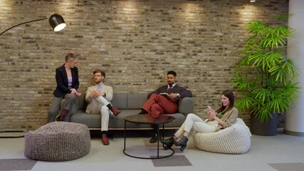 Businesspeople relax and use smartphones on a sofa and a beanbag chair in a modern office lobby with a brick wall and plants in the background
