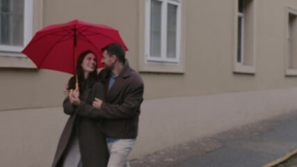 Romantic young couple walking closely under bright red umbrella, sharing tender moments while strolling through rainy urban street during autumn season - Powered by Adobe
