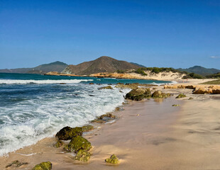 A stunning coastal view of a sandy beach on Margarita Island, Venezuela. Waves crash against mossy rocks while the shoreline stretches towards distant mountains under a clear blue sky.