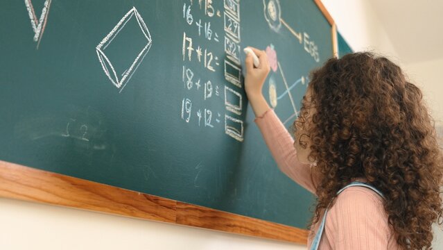 Back view of african student answer question while writing at board. Teacher looking at elementary student doing math quiz in front of classroom while smart children attend in math lesson. Pedagogy.