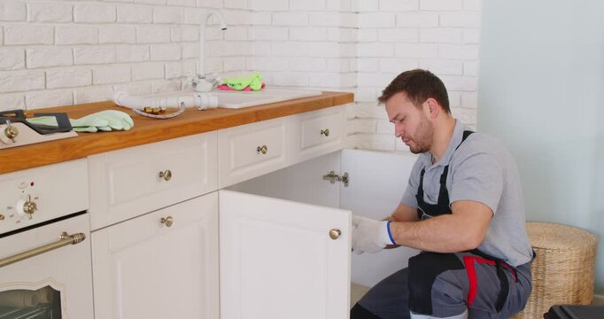 Male plumber in uniform kneeling on floor, fixing drain sink pipes inside kitchen cabinet. Professional repairman eliminating leaks or blockage, using tools for plumbing service and repair works.