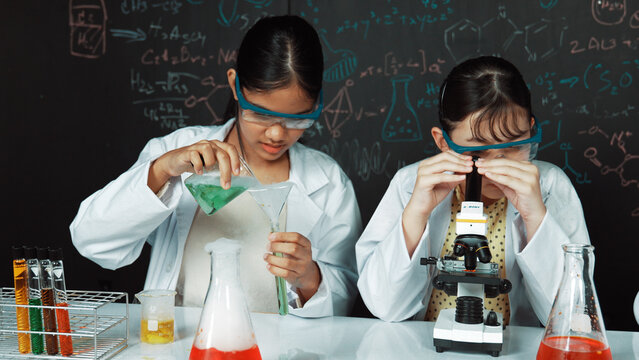 Young highschool student doing experiment by pouring sample in test tube. Girl wearing glasses and looking under microscope to inspect sample in STEM science class. Creative education. Edification.