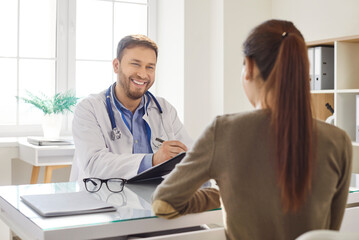 Young happy friendly male doctor consulting his woman patient sitting on workplace in examination...