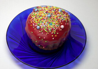 Easter cake, muffin with dark red melted sugar glaze and pastry nonpareil. colored sugar balls, scattered on sweet topping, on blue semitransparent elegance glass plate