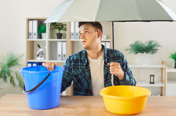 Dissatisfied man deals with dripping water from ceiling or roof leak at home, using bucket and...